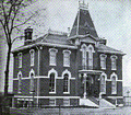 Black and white photo of a two-story brick building with ornate windows, a steep tower, and decorative trim, surrounded by a bare tree and a fence in the foreground.