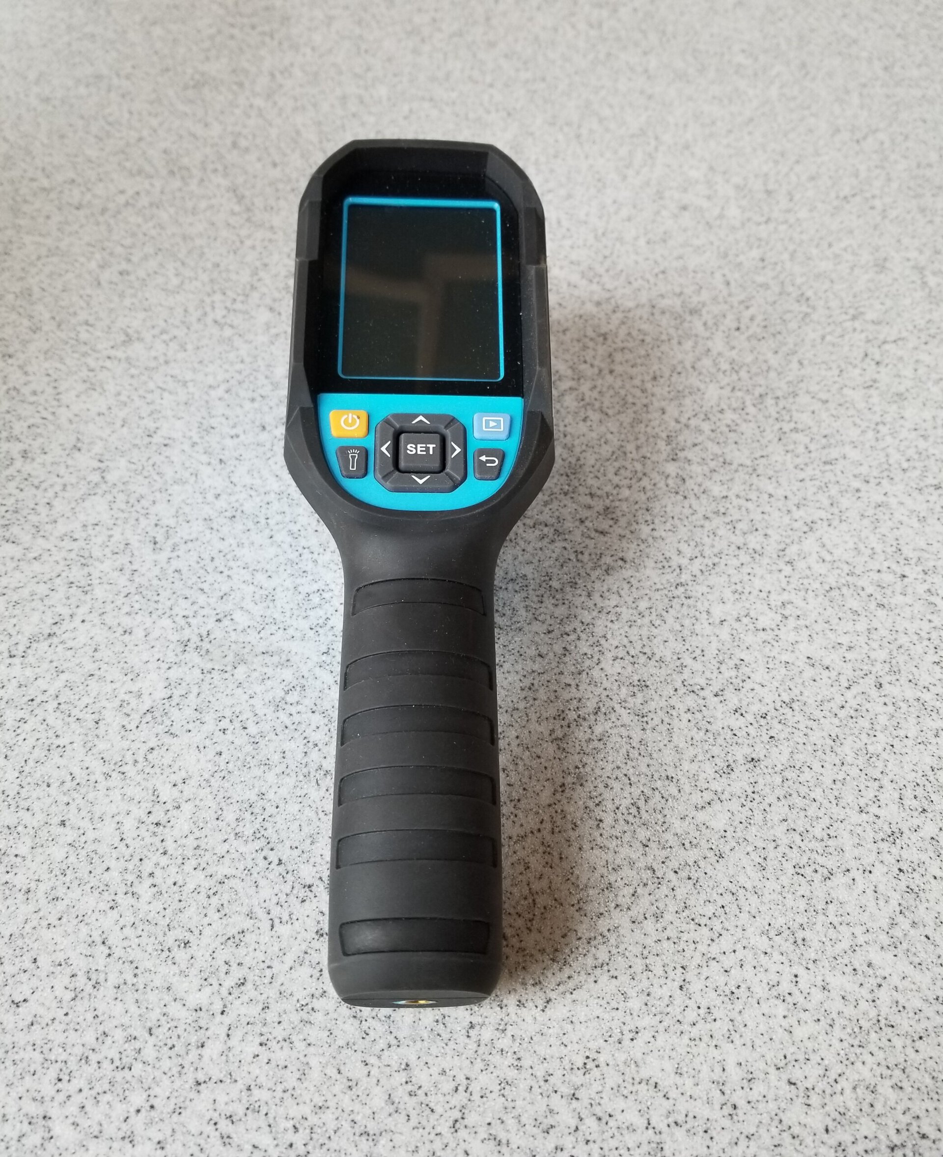 A handheld digital device with a screen, control buttons, and a black textured grip is resting on a gray speckled countertop.