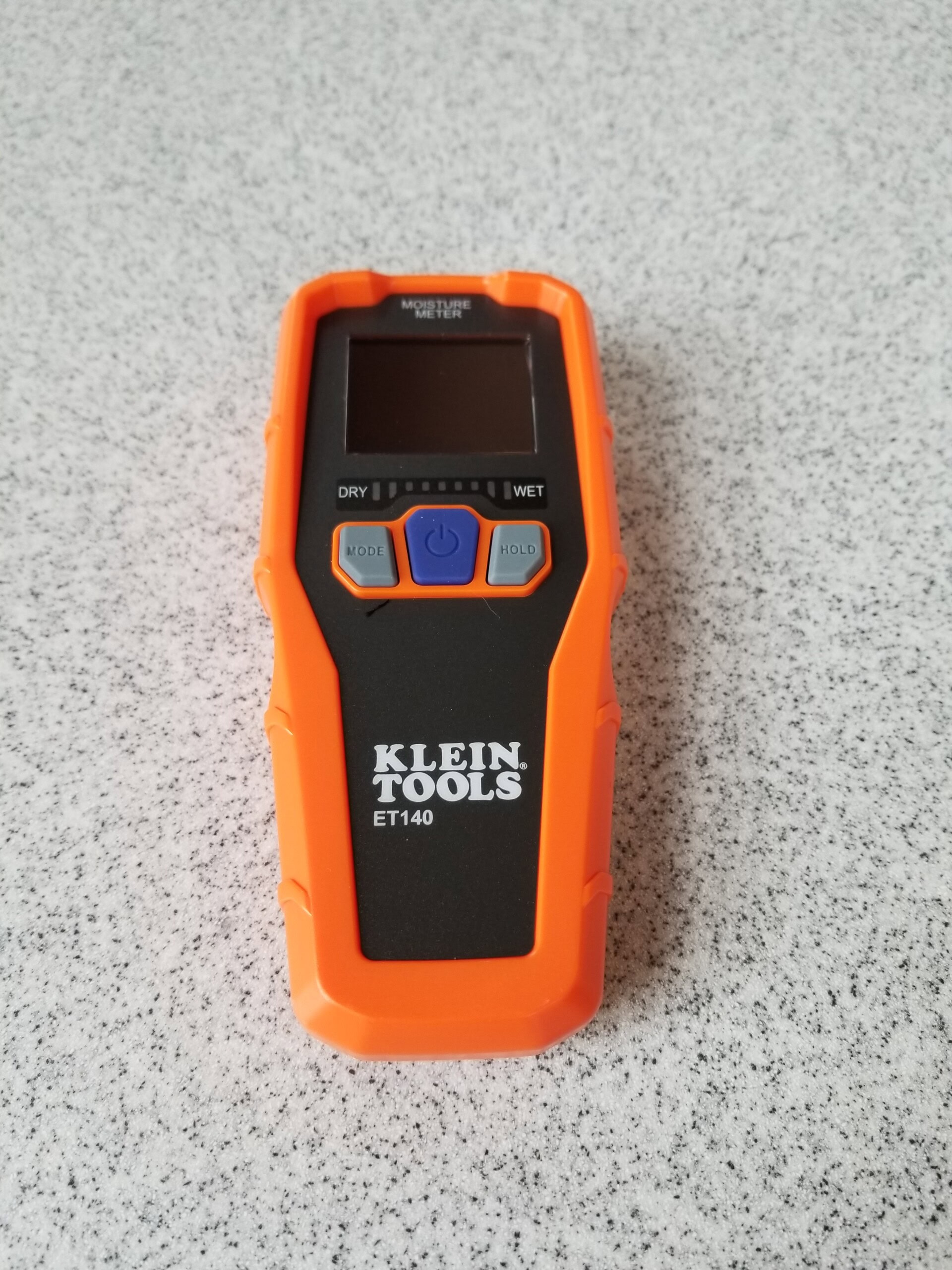A Klein Tools ET140 moisture meter with an orange case is placed on a light gray speckled surface. The device features a screen, buttons labeled MODE, HOLD, and a central blue power button.