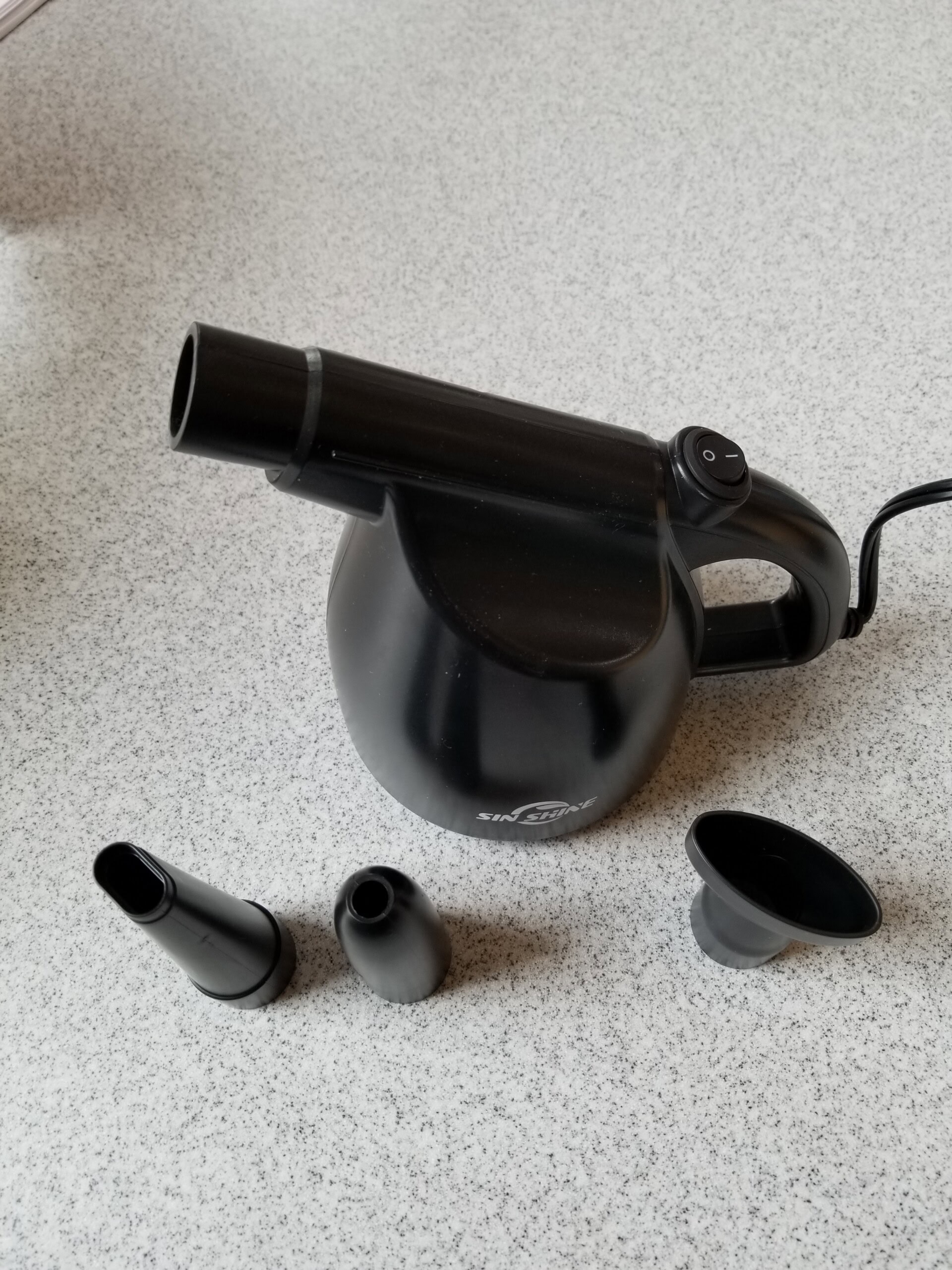 A black handheld steam cleaner with three different nozzle attachments is placed on a speckled countertop. One nozzle is attached to the device, while the other two are in front of it.