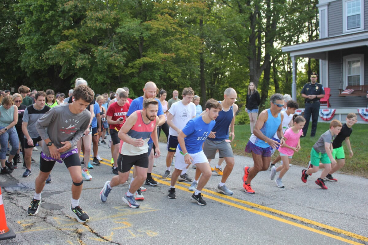 A group of runners of various ages line up and start a race on a street lined with trees, with spectators and a police officer watching from the sidewalk and a house porch in the background.